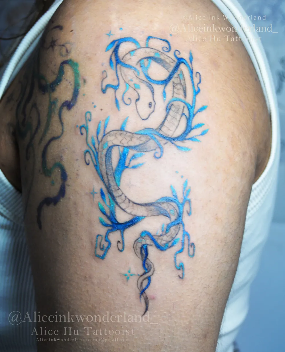 Blue fine-line snake tattoo winding across a shoulder, by Alice Ink Wonderland